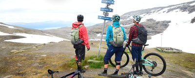 Mountain bikers wearing EVOC backpacks gathered around directional trail marker