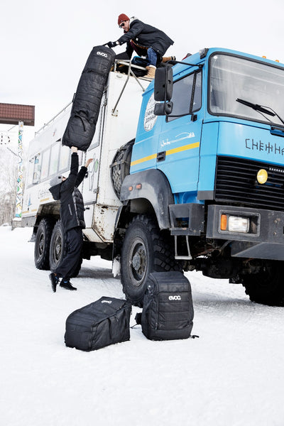 Two people unloading EVOC gear bags and ski travel roller bag from atop arctic transport truck in cold snow