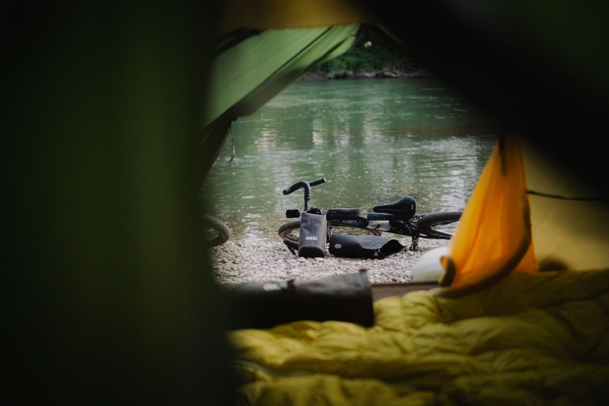 EVOC bikepacking bags and gear beside a river campsite at dusk, viewed from inside a tent