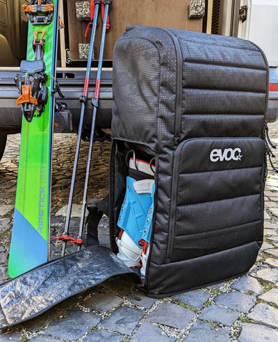 EVOC Gear Backpack with alpine ski touring boots showing in unzipped side compartment with skis a poles leaning next to it